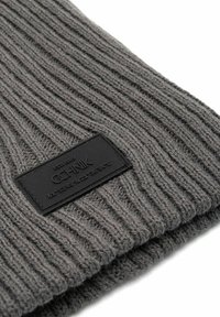 Szary, dzianinowy czapka z prążkowanym teksturą, z czarną skórzaną metką z nazwą marki i współrzędnymi. Miękki, elastyczny materiał.