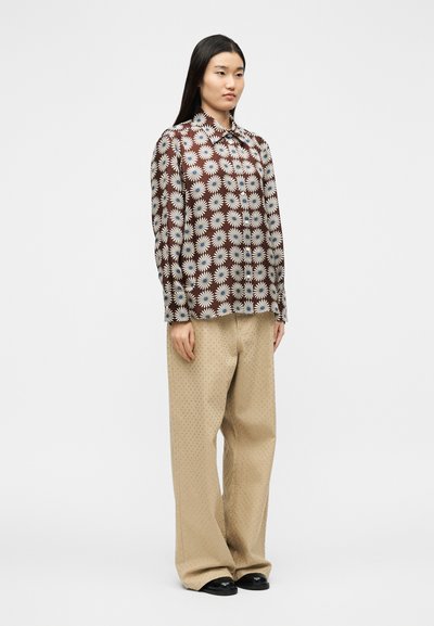Γυναίκ α που στέκεται, φορώντας καφέ μπλούζα μακρυμάνικη με λευκά σχέδια μαργαρίτας και μπεζ παντελόνι φαρδύ με διακριτική υφή πουά.