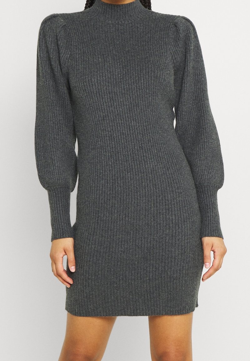 Femme portant une robe pull en maille côtelée gris foncé avec de longues manches bouffantes et un col montant, debout devant un fond clair uni.