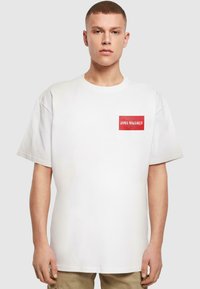 White cotton T-shirt with short sleeves, featuring a red rectangular patch with "DIMA MAGHREB" text and a star design. Relaxed fit.