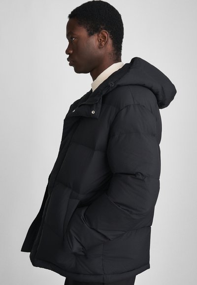 Jeune homme de profil portant une veste à capuche noire rembourrée avec les mains dans les poches, sur un fond uni.