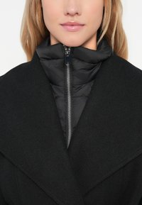 Cappotto nero con ampi revers, sovrapposto a un colletto interno imbottito con zip. Tessuto liscio con un design elegante e moderno.