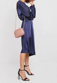 Abito di raso blu navy con orlo asimmetrico, abbinato a sandali neri con cinturini e una borsa a tracolla in oro rosa con texture intrecciata.
