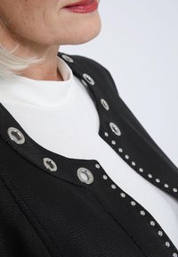 Veste noire texturée avec des œillets métalliques et des clous le long du col, portée sur un col roulé blanc. Gros plan sur l'encolure.