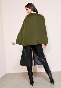 Oliivroheline kuub, millel on struktureeritud disain, koos mustade kunstnahast laiade pükste ja põlvikute tipuga saapastega, aksessuaariks sõrmus.