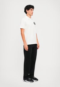 Νεαρός άνδρας με σγουρά μαλλιά, φορώντας λευκή μπλούζα polo με λογότυπο, μαύρο παντελόνι και μαύρα αθλητικά παπούτσια, που στέκεται μπροστά σε λευκό φόντο.