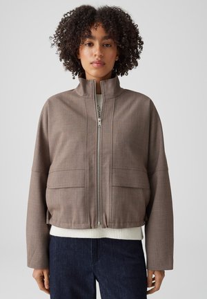 Femme aux cheveux bouclés portant une veste taupe zippée sur un pull blanc et un pantalon bleu foncé, debout devant un fond uni.