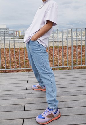 Person lehnt sich zurück gegen ein Geländer, trägt ein hellviolettes übergroßes T-Shirt, hellblaue Jeans und lila-orange Turnschuhe.