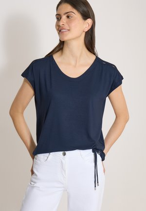 Femme portant un haut bleu marine à manches courtes avec un cordon et un pantalon blanc, debout les mains dans le dos, regardant vers la gauche.