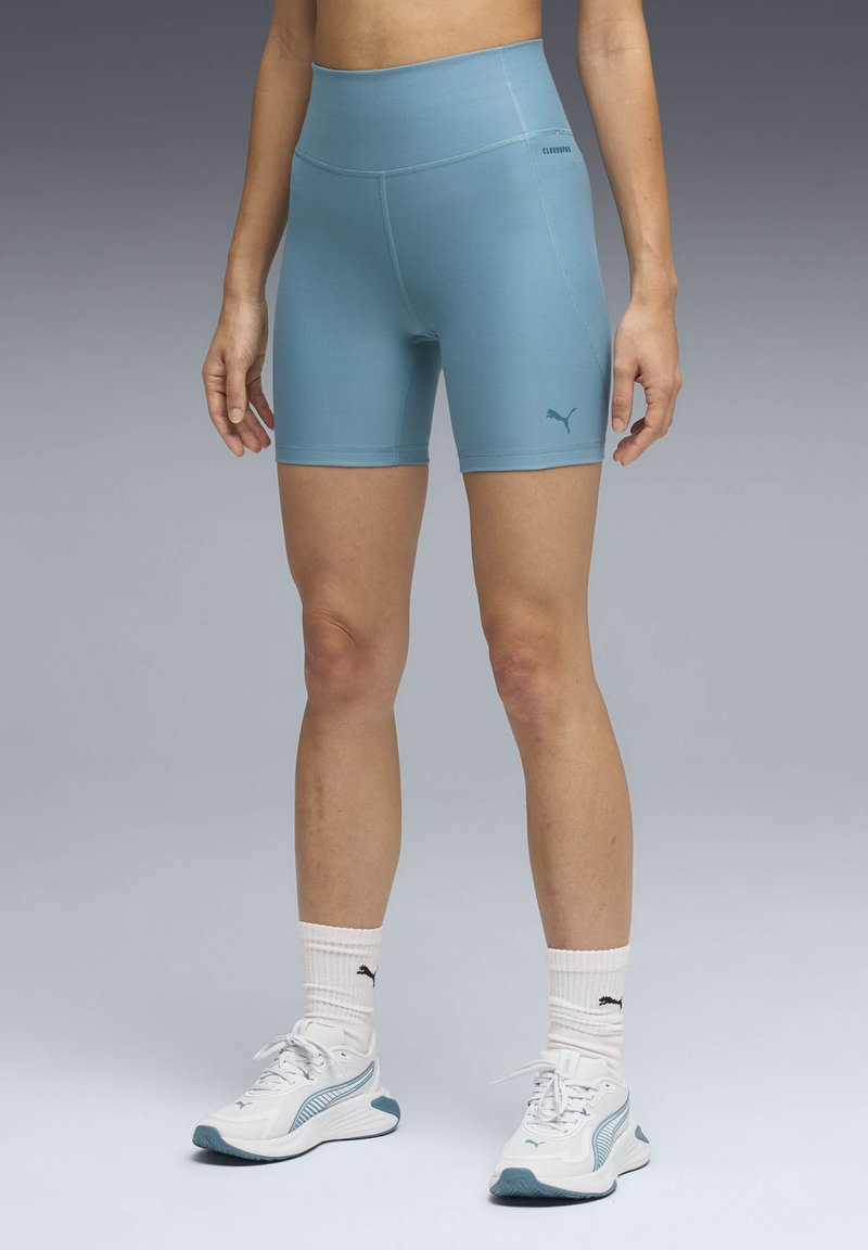 Άτομο που φοράει ανοιχτό μπλε ποδηλατικά σορτς Puma, λευκές κάλτσες Puma και λευκά και μπλε αθλητικά παπούτσια Puma, στέκεται σε γκρι φόντο.