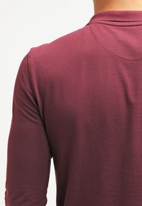 Camisa polo de manga larga color burdeos con tejido texturizado, cuello y un diseño ajustado. Detalles de costura visibles en los hombros y las mangas.