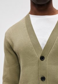Olivovo zelený žebrovaný kardigán s V-výstrihom, ozdobený tromi čiernymi gombíkmi a prehodený cez biele tričko. Štruktúrovaná pletenina.