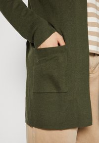 Cardigan verde scuro con trama a costine, dotato di tasca frontale e maniche lunghe. Indossato sopra una camicia a righe e pantaloni di colore chiaro.