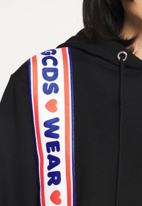 Sweat à capuche noir avec une sangle d'épaule colorée et graphique portant les textes "GCDS" et "WEAR" en bleu et rouge sur fond blanc.