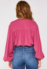 Blusa lunga a maniche lunghe in tessuto rosa, morbida e ampia, con dettaglio arricciato sul retro, abbinata a jeans a vita alta blu. Texture liscia e design rilassato.