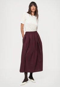 Haut à manches courtes à rayures blanches associé à une longue jupe bordeaux à plis ; le mannequin porte des collants noirs et des chaussures blanches à petit talon.