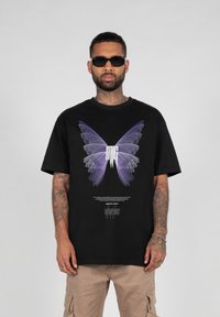 Sort oversized t-shirt med et lilla sommerfuglegrafik og tekstdesign på fronten. Stoffet ser blødt ud med en afslappet pasform.