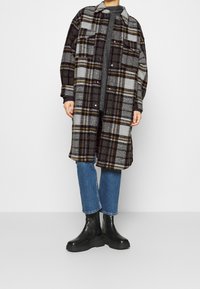 Cappotto a quadri in grigio, nero e marrone, con vestibilità rilassata, due tasche sul petto e bottoni a pressione. Abbinato a jeans blu e stivali neri.
