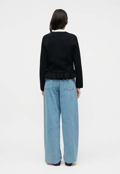 Γυναίκα με μακριά σκούρα μαλλιά που φορεί μαύρο πουλόβερ με βολάν στο τελείωμα, ανοιχτό μπλε τζιν με φαρδύ πόδι και μαύρα παπούτσια, στέκεται με την πλάτη στραμμένη.