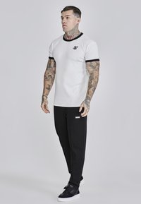 Witte T-shirt met korte mouwen en zwarte ribbelaccenten, gecombineerd met zwarte joggingbroek met een logo-patch, en zwarte sneakers.