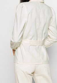 Chaqueta beige clara con cintura ceñida por un cinturón, mangas abullonadas y cuello con solapa, hecha de una tela suave con una textura lisa.