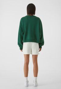 Zelená oversized mikina s žebrovanými manžetami a lemem, spárovaná s bílými šortkami. Model má na sobě bílé tenisky a žebrované ponožky do půli lýtek.