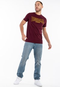 Burgundi T-shirt su geltonu "Lonsdale London" logotipu, trumpomis rankovėmis, derinama su šviesiai mėlynomis džinsinėmis kelnėmis ir baltais sportbačiais, stovint prieš baltą fono.