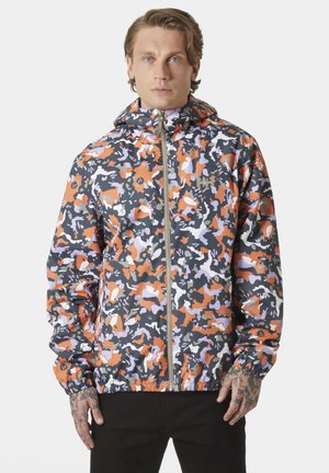 Mężczyzna w wielokolorowej kurtce z kapturem na zamek w kolorach pomarańczowym, białym, czarnym i szarym moro, z elastycznymi mankietami, stojący na jednolitym tle.