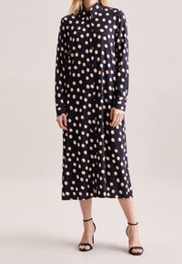 Largo vestido azul marino con grandes lunares crema, botón frontal, cuello con solapa y mangas largas. Llevado con tacones negros de tiras.