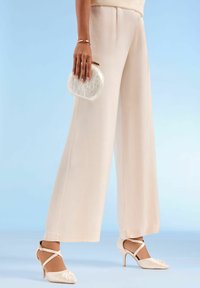 Hlače širokih hlačnic, kremne barve z gladko teksturo; kombinirane z natikači kremne barve z detajlom vozla in prozorno torbico.
