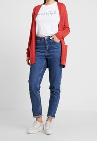Röd stickad cardigan, vit grafisk t-shirt, blå högmidjade jeans och vita sneakers. Cardiganen har öppen framsida och sidofickor.