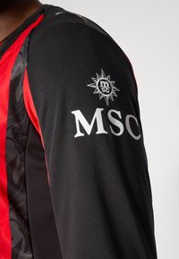 Sort og rød sportstrøje med tekstureret stof, der har et fremtrædende "MSC" logo og et cirkulært emblem på ærmet.
