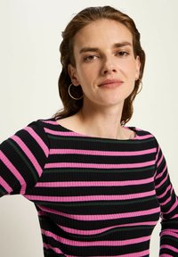 Frau mit hellbraunem Haar, die goldene Creolen trägt und ein schwarz, pink und grün gestreiftes Oberteil in horizontaler Ausrichtung anhat, schaut frontal vor einem einfarbigen Hintergrund.