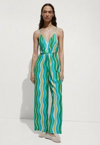 Tuta a fantasia verde, blu e arancione con strisce ondulate. Presenta spalline sottili e un scollo profondo, con vita aderente e gambe larghe.
