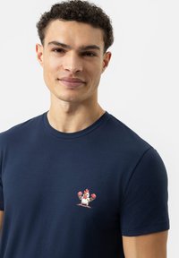 Jonge man met kort, krullend haar die een marineblauwe t-shirt draagt met daarop een kleine cartoonkip in rode bokshandschoenen.