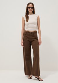Mujer con top beige sin mangas, pantalones marrones de pierna ancha, sandalias negras con tacón, pendientes de aro y gafas de sol rectangulares oscuras, de pie frente a un fondo liso.