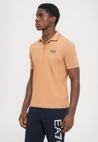 Brun polo-skjorte med krage, to knapper og brodert "EA7"-logo. Kombinert med marineblå bukser med hvit tekstdesign.