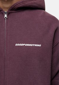 Hoodie bordeaux avec fermeture éclair, arborant le texte blanc "GOODFORNOTHING" sur la poitrine gauche. Fabriqué en tissu doux et texturé.