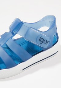 Sinine läbipainealne plastikust sandaal suletud varbaga, reguleeritava rihmaga, valge tald ja sile, tekstuuriga sisetald. Brändinimi "igor" nähtav.