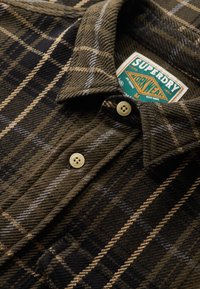 Camisa de franela a cuadros marrón con cuello abotonado, que presenta rayas beige y negras, botones redondos color crema y una etiqueta de marca visible.