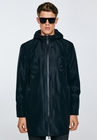 Jauns vīrietis, kurš valkā melnu lietussarga mēteļa kapuci ar rāvējslēdzēju un auklas kapuci, komplektā ar tumšām saulesbrillēm, stāv pret vienkāršu fonu.