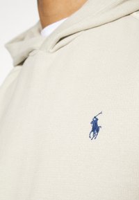 Sweat à capuche beige en tissu texturé, avec une poche avant et un logo brodé bleu représentant un cheval avec un cavalier sur la poitrine gauche.