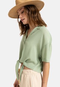 Blusa verde claro de manga corta con un tejido texturizado, cuello y detalle de nudo atado en la cintura. Combinada con pantalones sueltos de color beige.