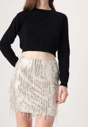 Woman wearing a black cropped long-sleeve top and a high-waisted beige sequin fringe mini skirt, posing with one hand on hip.