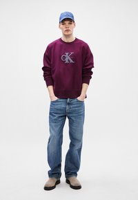 Burgundská mikina s vyšitým logem, volný střih, žebrované manžety, v kombinaci s modrými džíny a šedými nazouváky, doplněné světle modrou kšiltovkou.