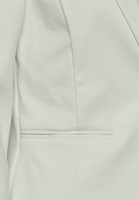 Gros plan d'une veste en tissu gris clair montrant une poche passepoilée et des lignes de couture sur un matériau texturé.