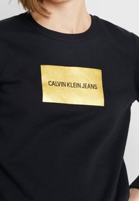 Czarna bluza o kroju swetra, wykonana z tkaniny bawełnianej z domieszką, z błyszczącym złotym prostokątnym logo, na którym widnieje napis "CALVIN KLEIN JEANS" dużymi czarnymi literami.