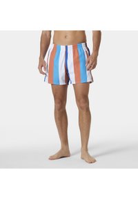 Mężczyzna stoi boso w kostiumie kąpielowym w pionowe szerokie paski w kolorach niebieskim, białym i rdzawego pomarańczowego na jednolitym tle.