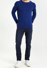 Blå långärmad tröja med rund halsringning, kombinerad med mörkblå slim-fit byxor och vita sneakers för en koordinerad avslappnad look.