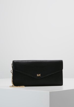 Portefeuille Michael Kors en cuir noir avec logo MK doré et chaîne dorée reposant sur une surface blanche sur un fond gris uni.